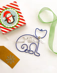 Personalized cat ornament, featuring an image of a cat outline in blue wire with the name Milo in the center in silver wire with white bow and silver bell. The Christmas wrapping paper, with a penguin design, lies diagonally, with part of a ribbon running diagonally to the right, the remaining wrapping paper in the upper left corner of the image. The light green ribbon is to the right and runs downward in a gentle arc, while a light beige tag with a snowflake design is below the wrapping 