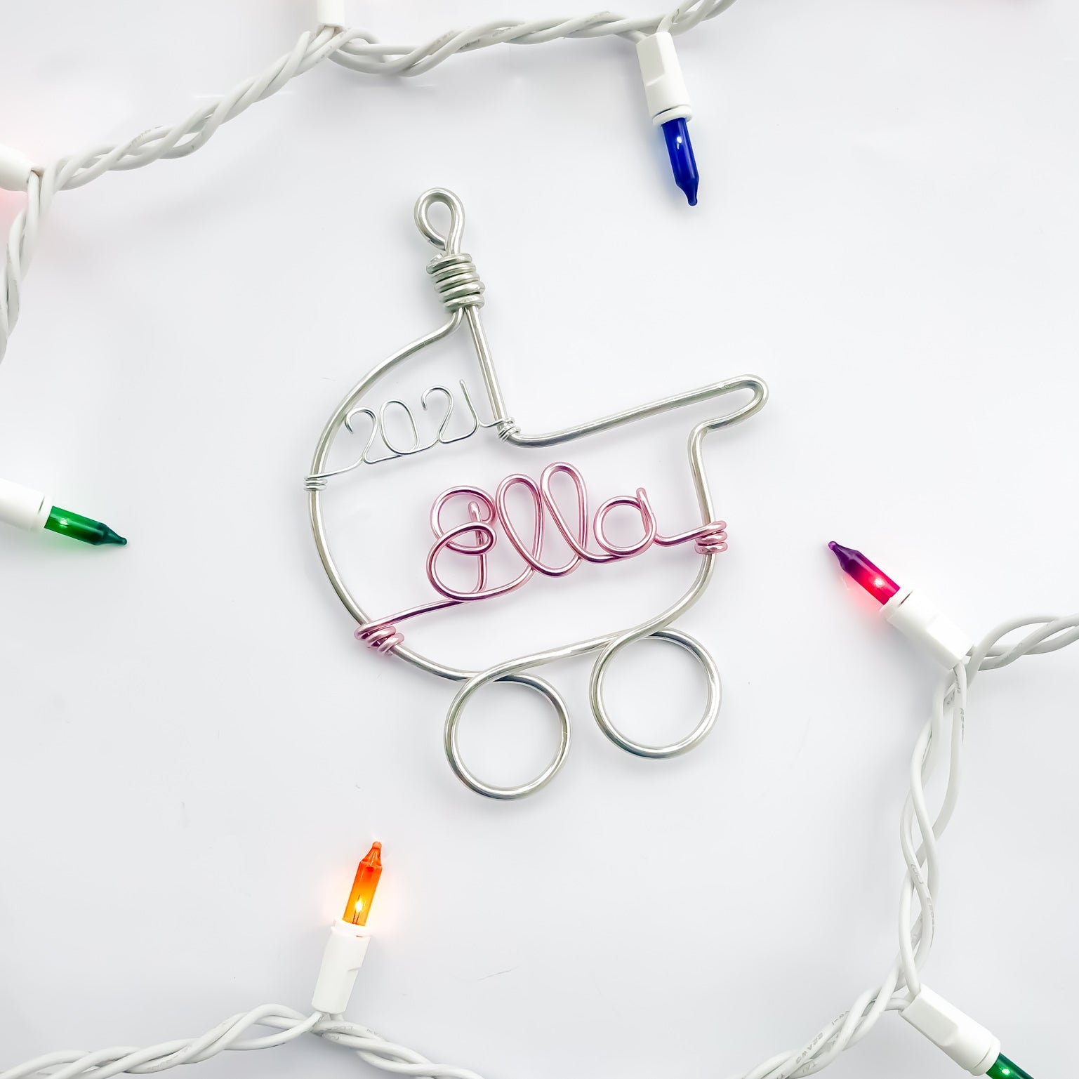 A wire-wrapped baby stroller ornament, featuring the name "Ella" and the year "2021", is nestled among a string of colorful Christmas lights.