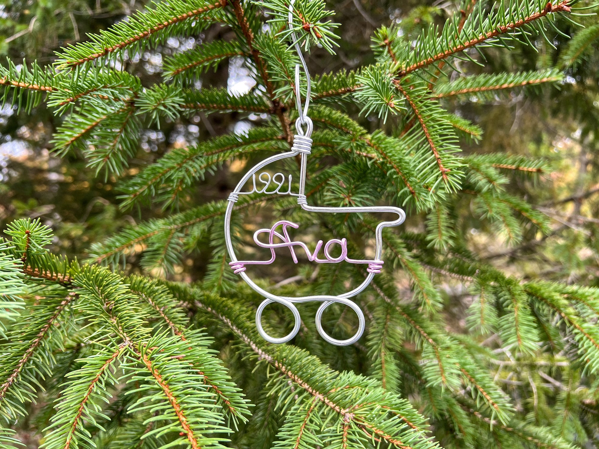 The image shows a wire-wrapped baby stroller ornament, one in silver aluminum featuring the name "Ava" in light pink and the year "1991,"  all set against a Christmas tree background.