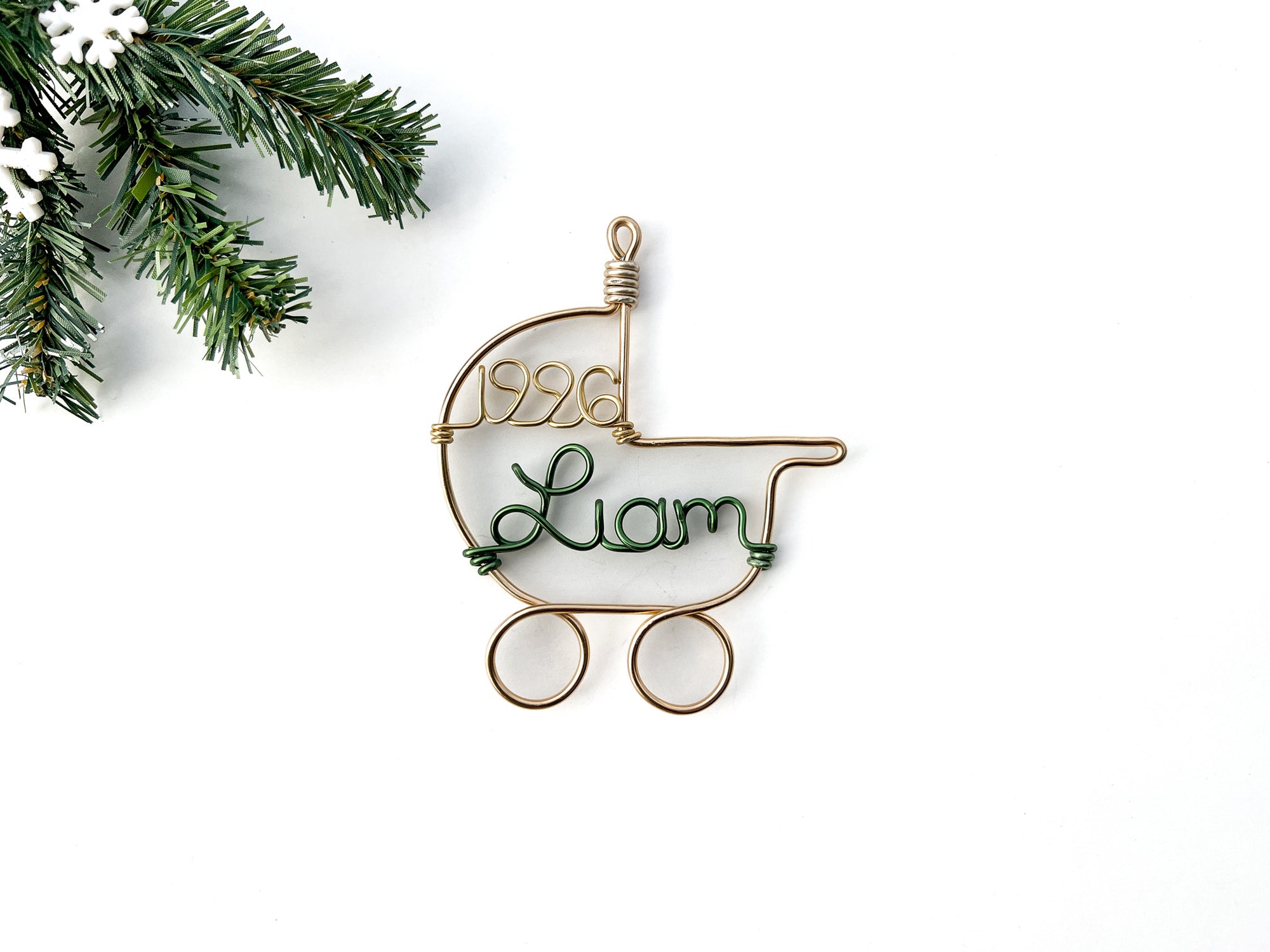 A wire-wrapped baby stroller ornament in gold aluminum, featuring the name "Liam" in green  and the year "1996" in gold, in a white background with a christmas tree branch in the corner.