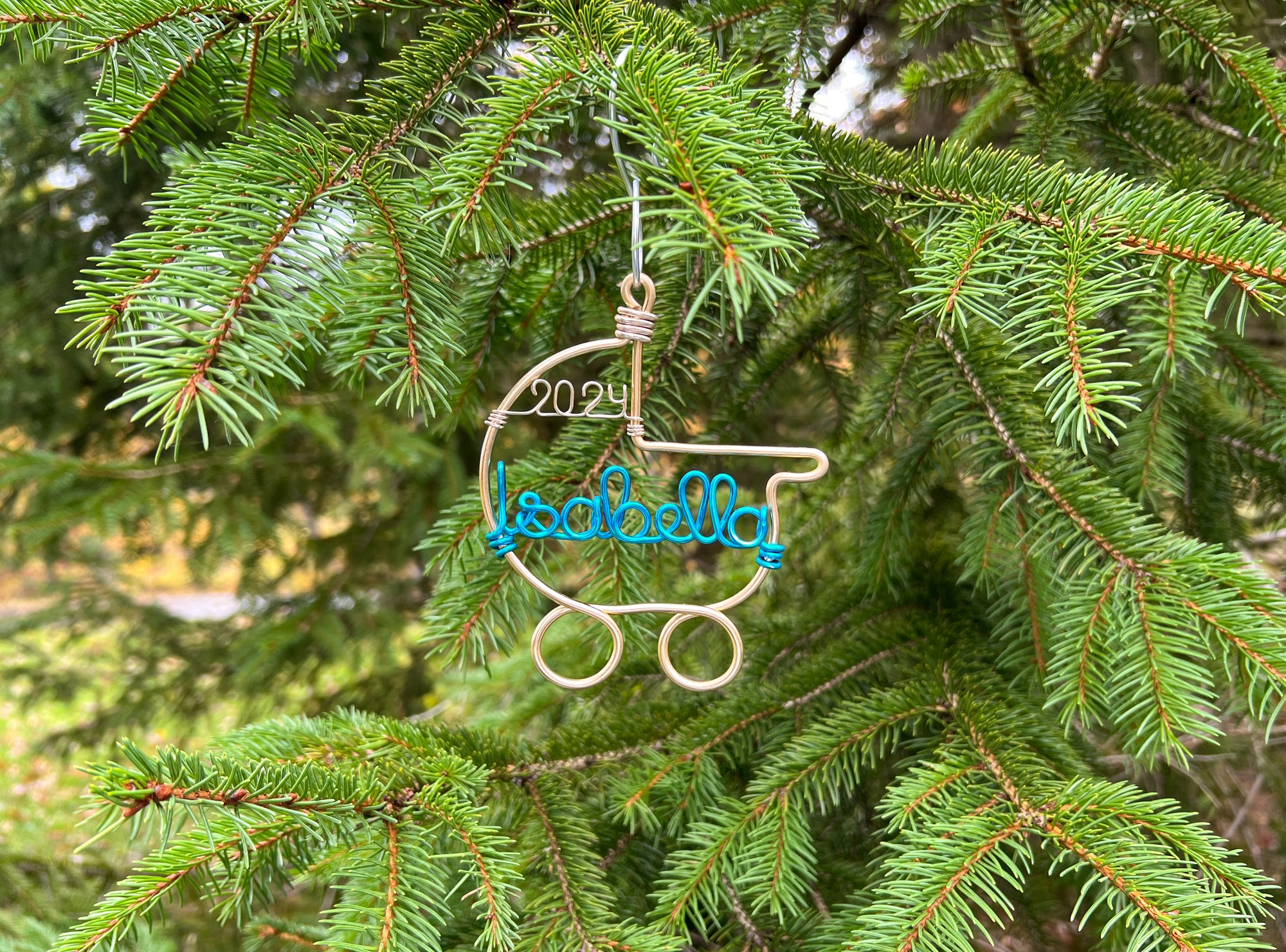 A wire-wrapped baby stroller ornament in gold aluminum, featuring the name "Isabella" in turquoise and the year "2024" in gold, is nestled among a Christmas tree hung by a silver hook.