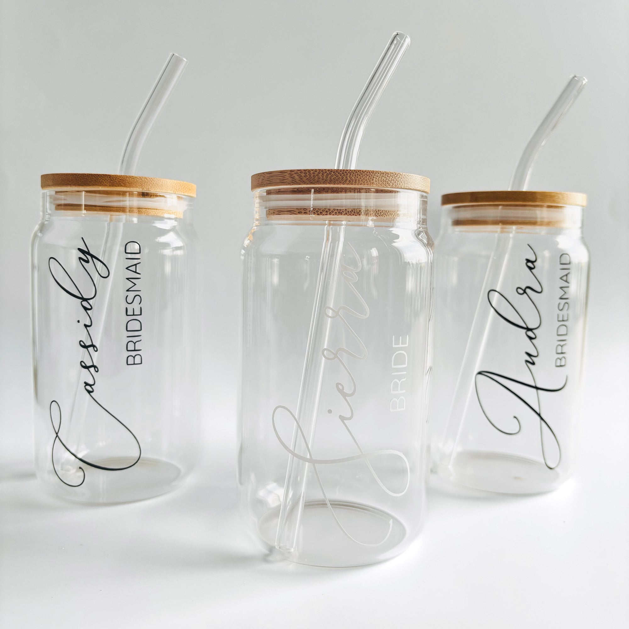 The image features three personalized glass straw tumblers, each with a cylindrical shape, bamboo lid, and glass straw. From left to right, they are personalized with the names and titles: Cassidy, Bridesmaid in black lettering; Sierra, Bride in the center in white lettering; and Andra, Bridesmaid on black lettering on the right, all set against a white background.