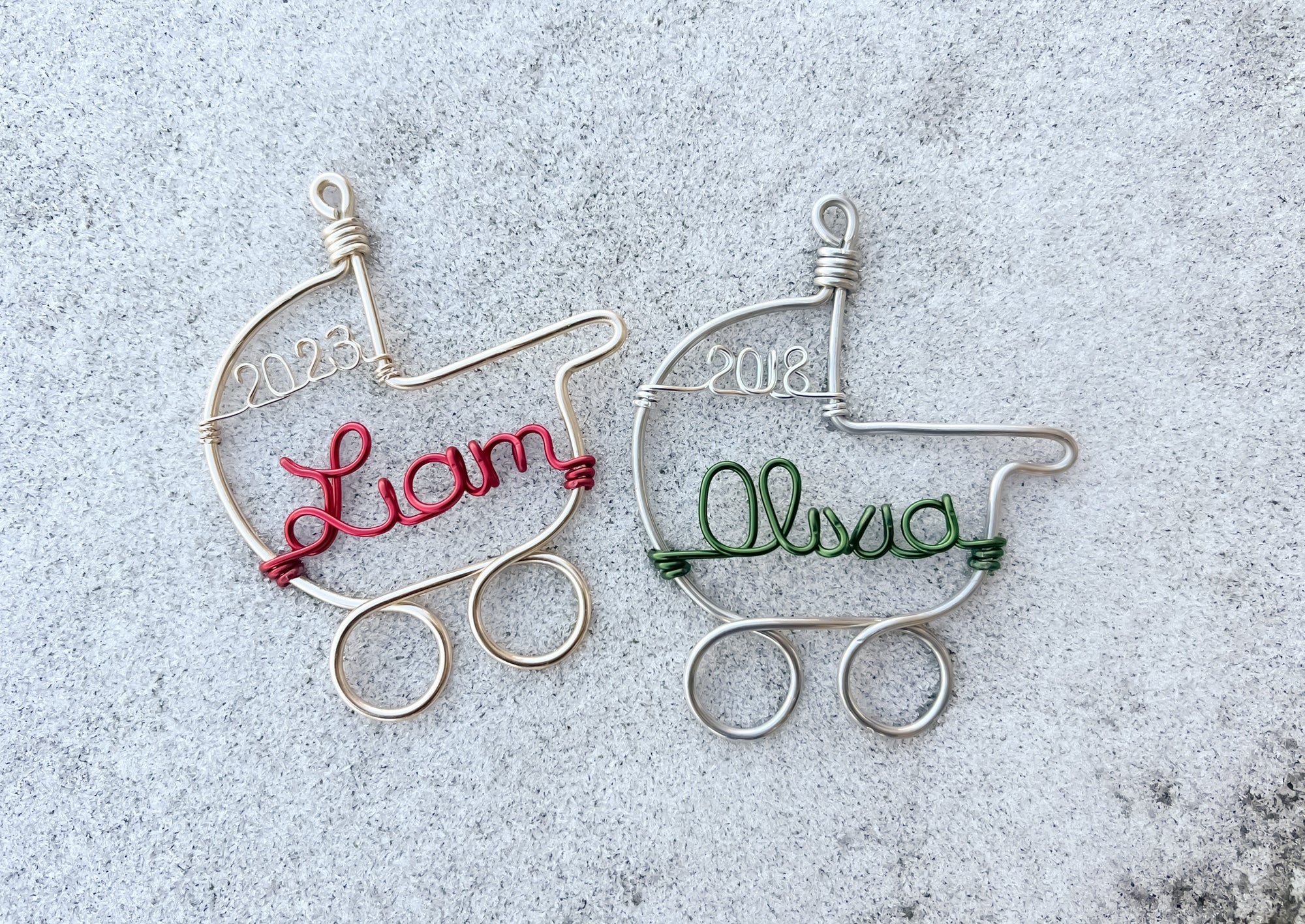 The image shows two wire-wrapped baby stroller ornaments, one in gold aluminum and the other in silver. The gold ornament features the name "Liam" in red and the year "2023," while the silver ornament features the name "Alissa" in green and the year "2018," all set against a gray background.