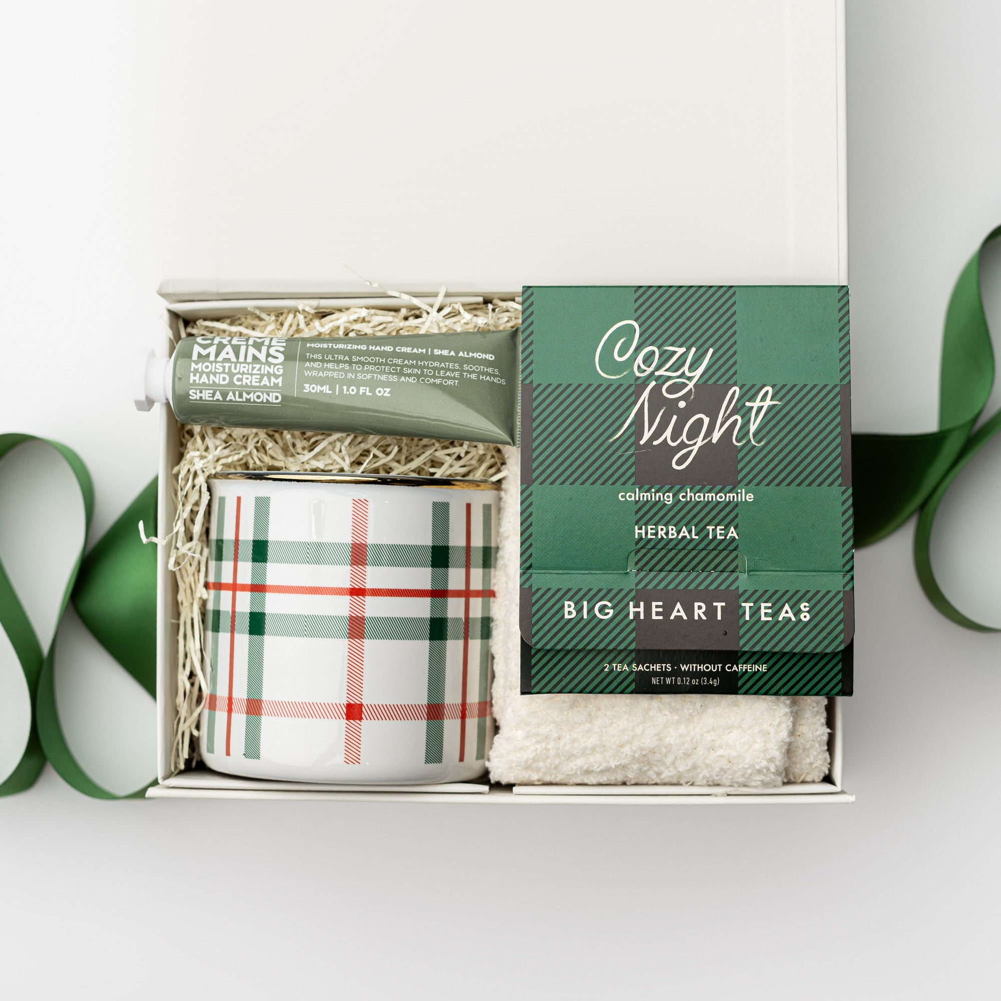 Petite cozy at home gift box featured in a small white box with ivory shred in the back with green ribbon crossing under it. Inside of the box on the left side almond and shea butter hand cream in its green tube container and plaid ceramic mug with white and green lines. On the right cozy night herbal tea in its green and black  plaid pattern package and underneath white cozy socks. All on a white background. 