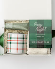 Petite cozy at home gift box featured in a small white box with ivory shred in the back with green ribbon crossing under it. Inside of the box on the left side almond and shea butter hand cream in its green tube container and plaid ceramic mug with white and green lines. On the right cozy night herbal tea in its green and black  plaid pattern package and underneath white cozy socks. All on a white background. 