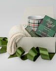 Petite Cozy at Home Holiday Gift is featured in a white box wrapped with a green ribbon. Inside the box, on the right, are white cozy socks; in the middle, a plaid mug with green and red lines; and on the left, a Cozy Night herbal tea in black and green plaid packaging. Everything is displayed on a white background.