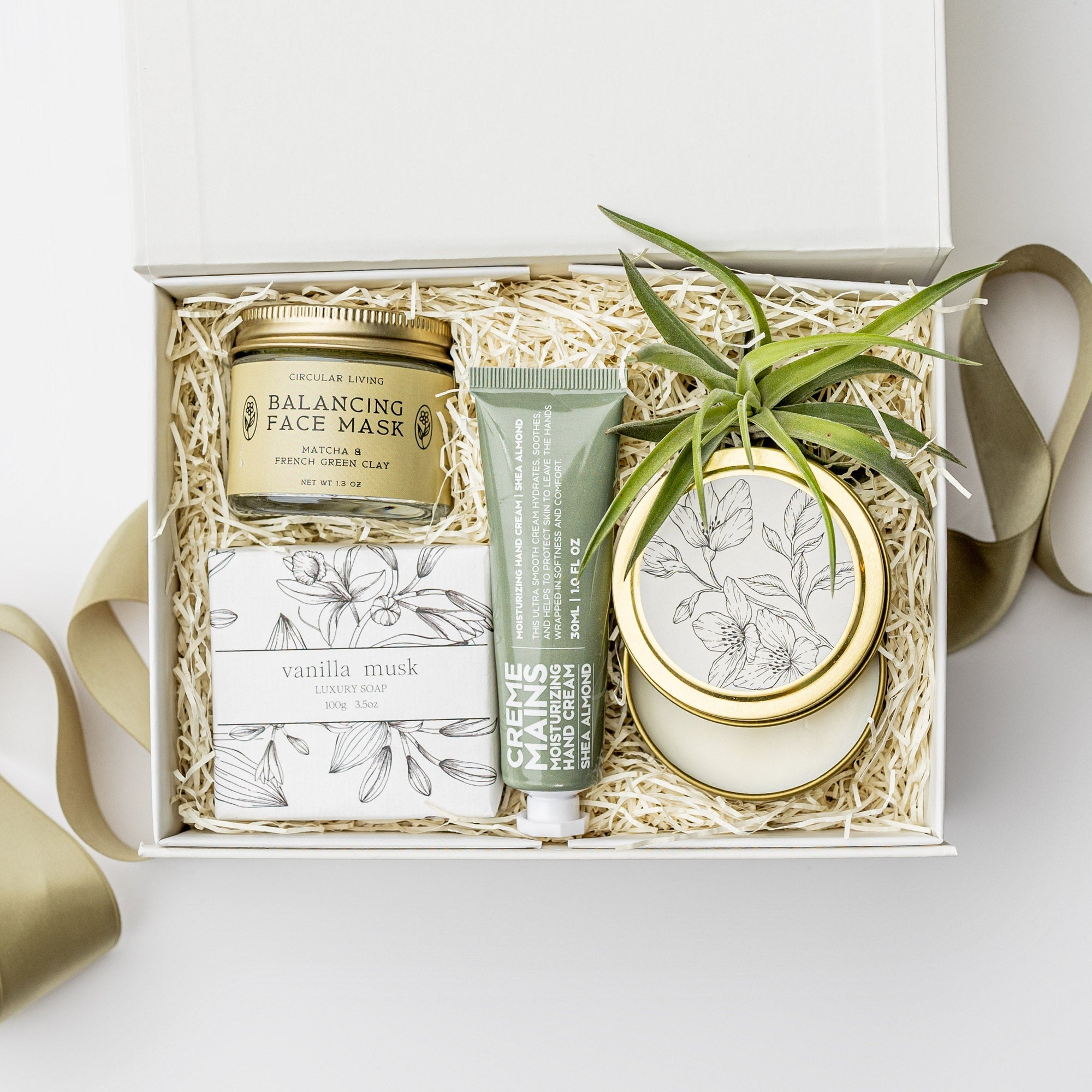 A white gift box with ivory paper shred and a green ribbon on a white background. Inside the box are a small live air plant, almond shea butter hand lotion, a vanilla musk bar soap, a matcha and green clay balancing face mask, and a 4oz hand-poured soy candle in the scent White Tea — a petite spa gift box designed for relaxation and self-care.