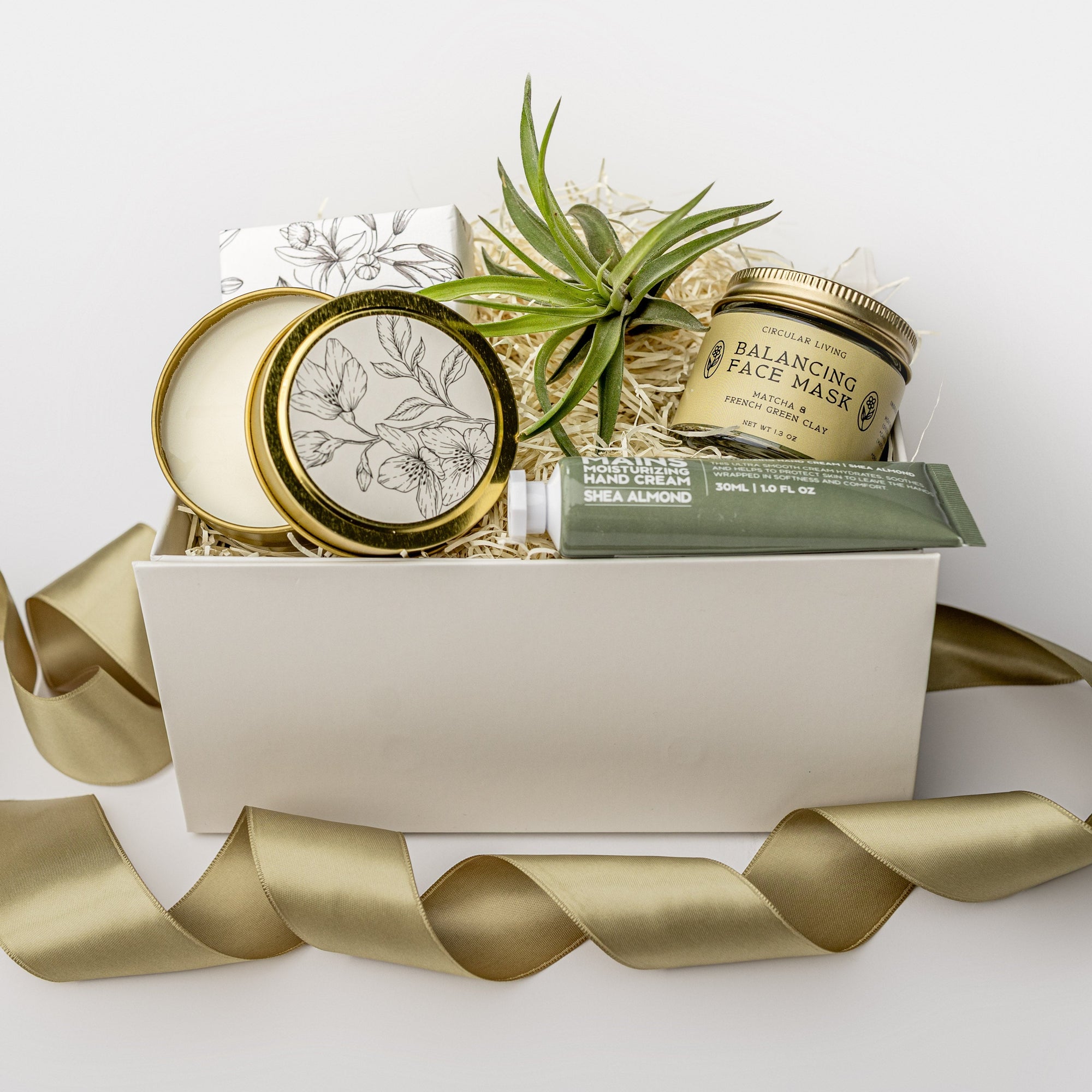 A white gift box with ivory paper shred and a green ribbon on a white background. Inside the box are a small live air plant, almond shea butter hand lotion, a vanilla musk bar soap, a matcha and green clay balancing face mask, and a 4oz hand-poured soy candle in the scent White Tea — a petite spa gift box designed for relaxation and self-care.