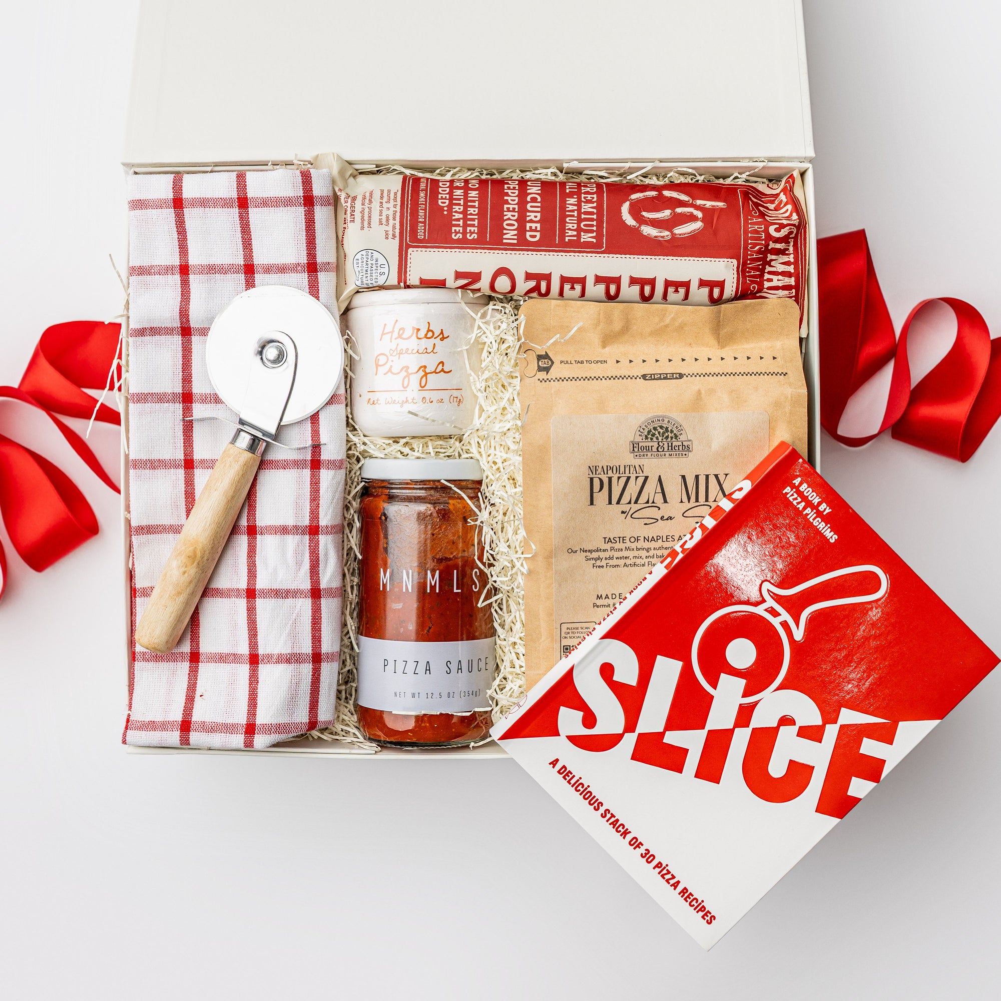 A white gift box with ivory paper shred and tied with a red ribbon on a white background. Inside the box are a Slice: Pizza Recipes book, a circular pizza cutter, a red grid tea towel, pizza herbs in a ceramic crock, an organic pizza flour mixture, an organic all-natural pizza sauce, and artisan all-natural uncured pepperoni — a “Pizza Night” gift box.