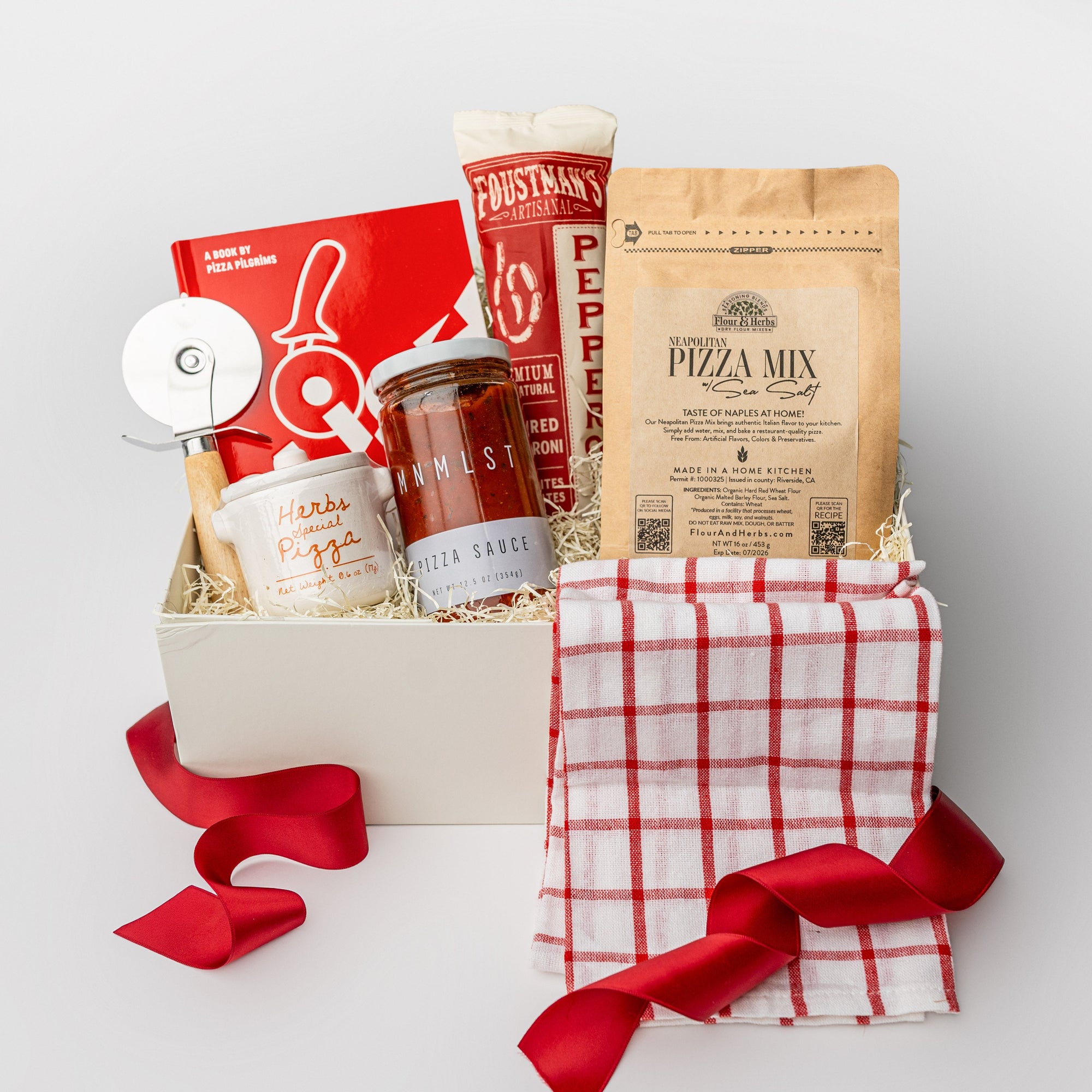 A white gift box with ivory paper shred and tied with a red ribbon on a white background. Inside the box are a Slice: Pizza Recipes book, a circular pizza cutter, a red grid tea towel, pizza herbs in a ceramic crock, an organic pizza flour mixture, an organic all-natural pizza sauce, and artisan all-natural uncured pepperoni — a “Pizza Night” gift box.