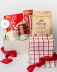 A white gift box with ivory paper shred and tied with a red ribbon on a white background. Inside the box are a Slice: Pizza Recipes book, a circular pizza cutter, a red grid tea towel, pizza herbs in a ceramic crock, an organic pizza flour mixture, an organic all-natural pizza sauce, and artisan all-natural uncured pepperoni — a “Pizza Night” gift box.