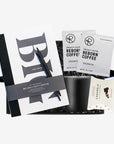 A curated gift box featuring a 12oz black vacuum-insulated camp mug with a handle, a large cream-paged Big Ideas sketchbook, individual pour-over coffee packs, a package of dark roasted mocha caramels, and a set of minimalist matte-black metal pens. All items are arranged inside a matte black Foxblossom. gift box filled with black shredded paper and tied with a black satin ribbon.