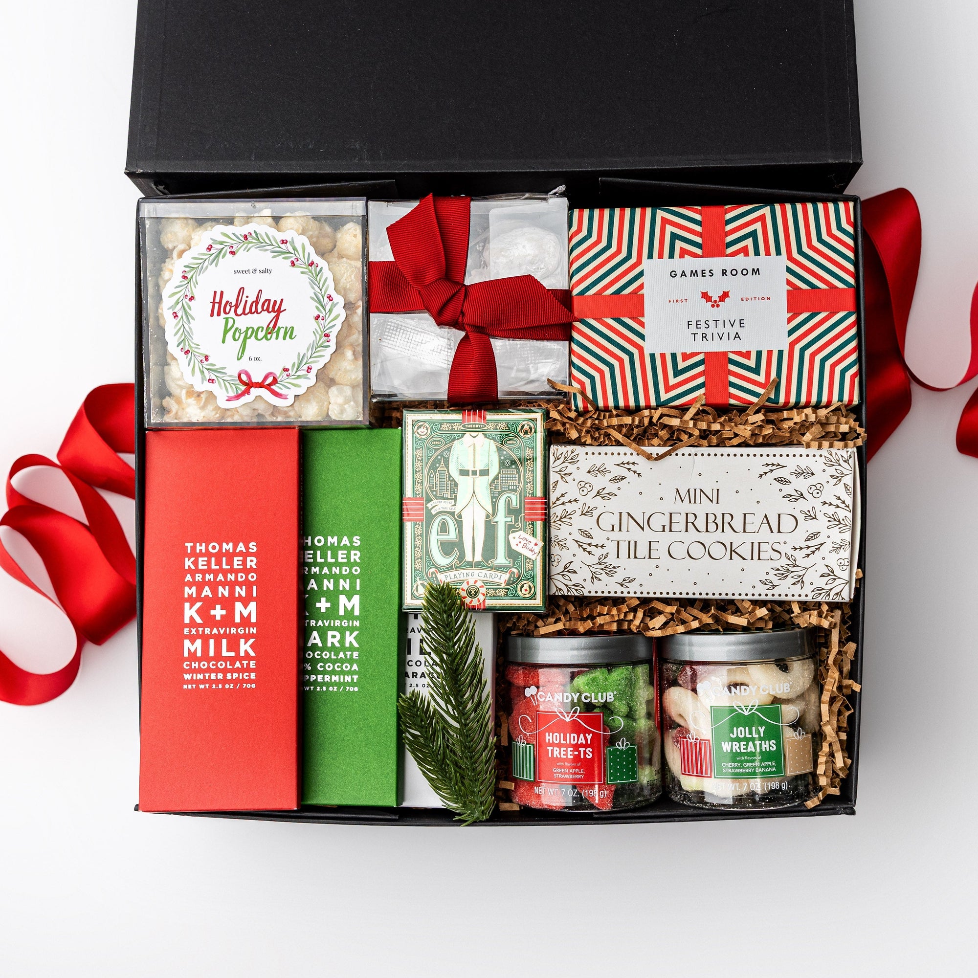 A black gift box with brown shredded paper inside and a red ribbon on a white background. Inside the box are sweet & salty popcorn, Lark Foods artisan tea cookies, a Ridley’s festive trivia game, Rustic Bakery mini gingerbread tile cookies, Jolly Wreaths gummies, holiday tree-shaped gummies, foil-stamped “Elf” luxury playing cards, and three K+M chocolate bars (winter spice, peppermint, and milk chocolate) — a “Reindeer Games” gift box ready for festive fun and treats.