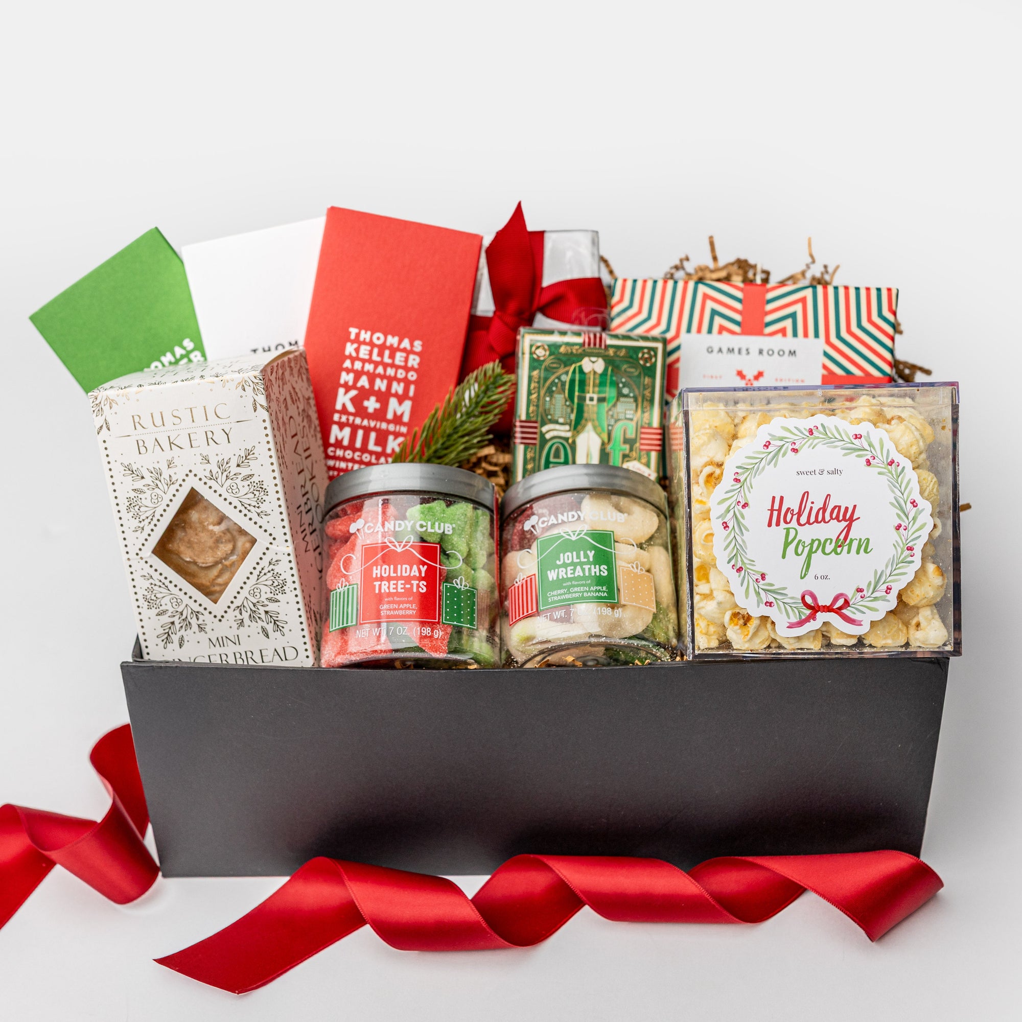 A black gift box with brown shredded paper inside and a red ribbon on a white background. Inside the box are sweet & salty popcorn, Lark Foods artisan tea cookies, a Ridley’s festive trivia game, Rustic Bakery mini gingerbread tile cookies, Jolly Wreaths gummies, holiday tree-shaped gummies, foil-stamped “Elf” luxury playing cards, and three K+M chocolate bars (winter spice, peppermint, and milk chocolate) — a “Reindeer Games” gift box ready for festive fun and treats.