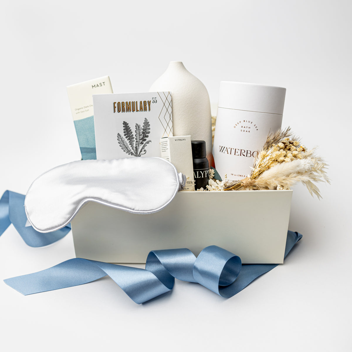 A curated wellness gift box set on a white background, featuring a white satin sleep mask, a white ceramic vase, skincare products from Waterbottle and Formulary, dried pampas grass, and a blue satin ribbon.
