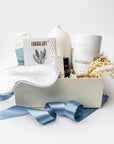 A curated wellness gift box set on a white background, featuring a white satin sleep mask, a white ceramic vase, skincare products from Waterbottle and Formulary, dried pampas grass, and a blue satin ribbon.
