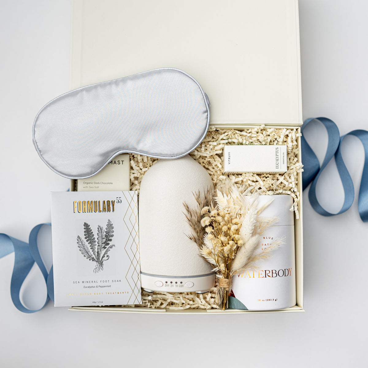 A curated wellness gift set arranged in an open cream-colored box with shredded paper filler. The set includes a light gray satin sleep mask, a white diffuser, a white candle, a boxed herbal tea, and dried pampas grass, with blue ribbons draped around the box.
