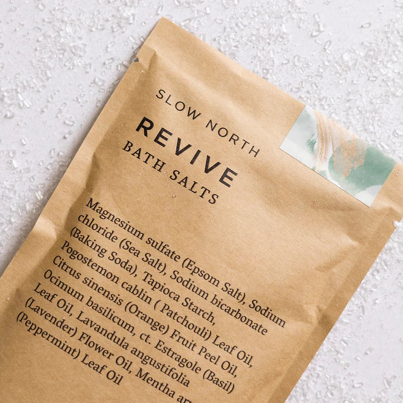 Slow North Revive bath salts in its brown packaging on a white background surrounded by the salts. 