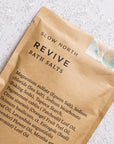 Slow North Revive bath salts in its brown packaging on a white background surrounded by the salts. 