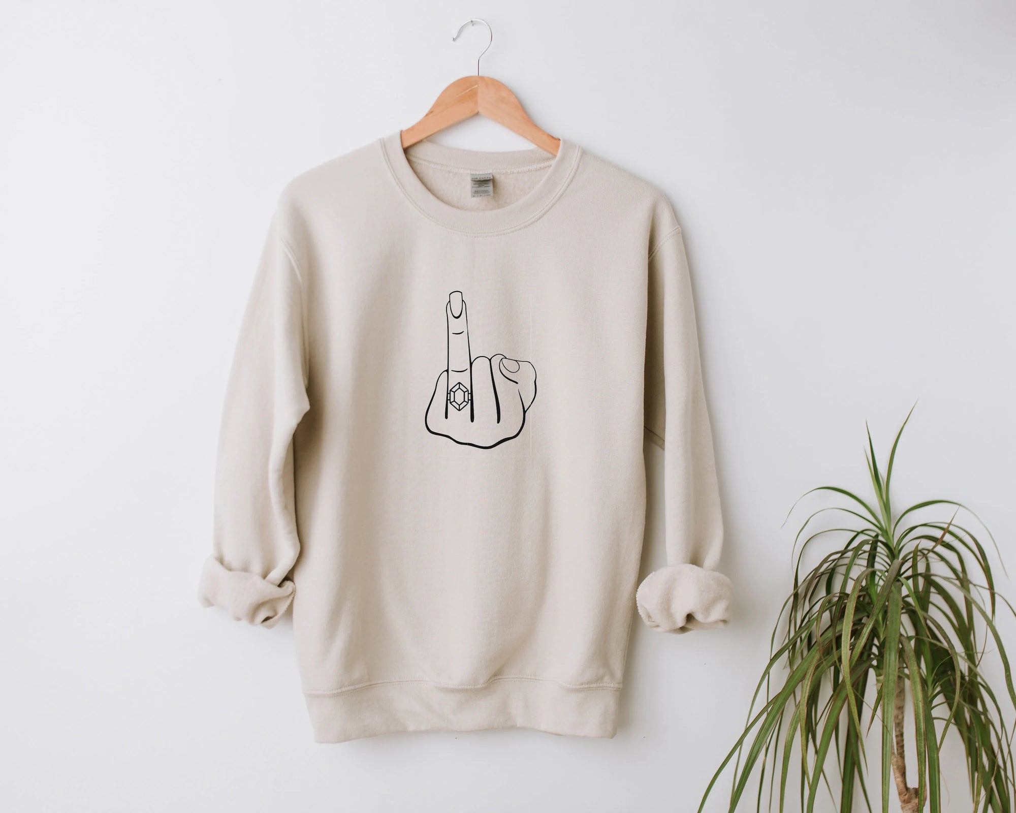 Ring finger engagement sweatshirt featured  that  has the image in black lines in the middle hanged on a white background with a plant on the right bottom corner.