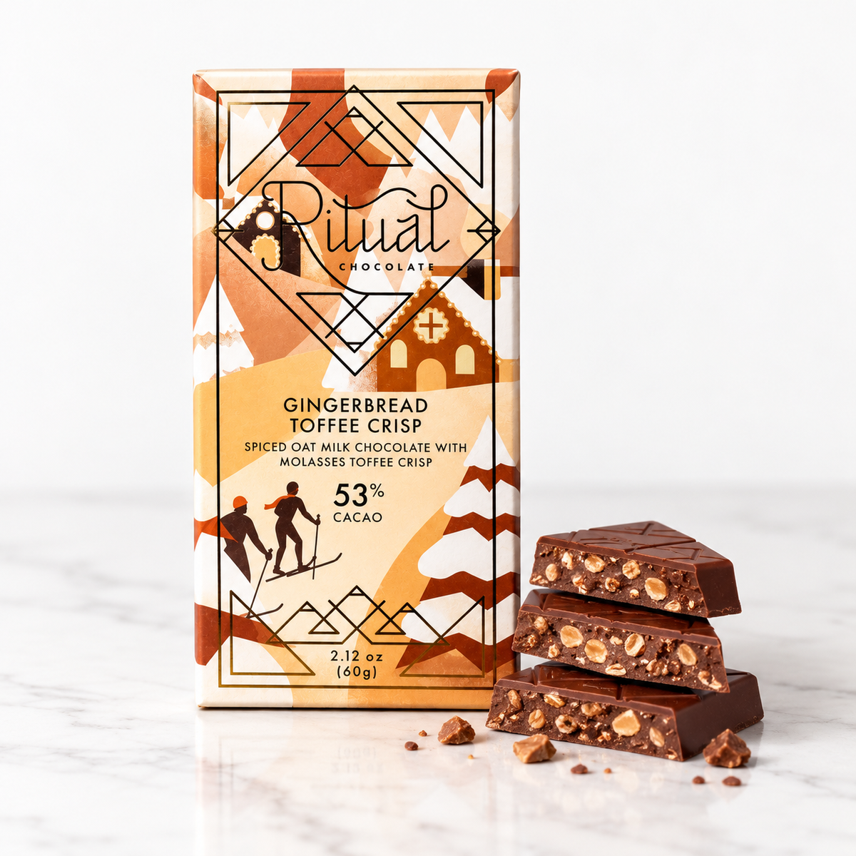 A package of Ritual Chocolate Gingerbread Toffee Crisp stands on a white marble surface with several broken chocolate pieces displayed in front. The box has an orange, beige, and brown geometric design with illustrations of skiers and houses.