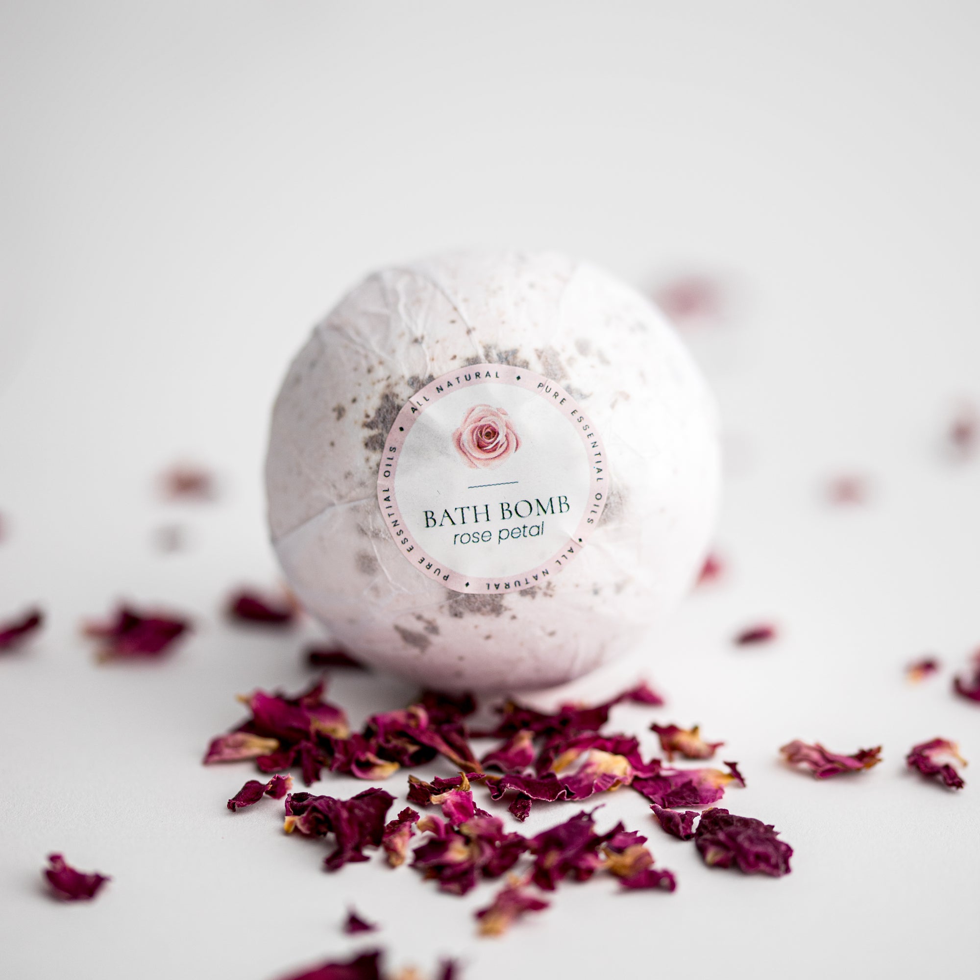 Rose Petal Bath Bomb, featured in its white transparent wrapping with a label that includes a rose illustration, pink details, and black lettering, surrounded by rose petals on a white background.