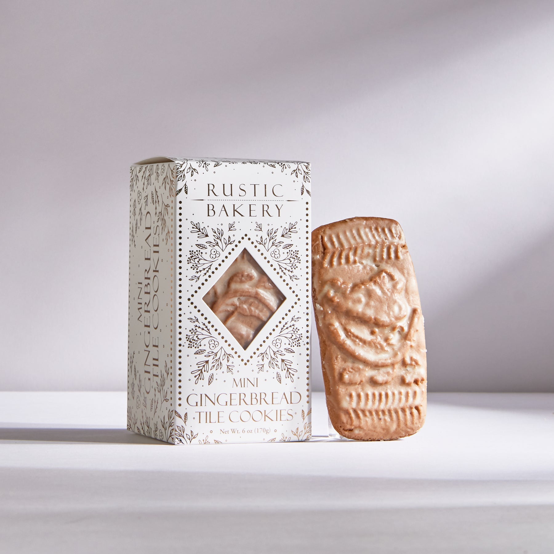 Rustic Bakery, Mini Gingerbread Tile Cookies, is featured in its white rectangular package with gold details and lettering. One of the square tile shaped cookies outside of the box next to it on a white background. 