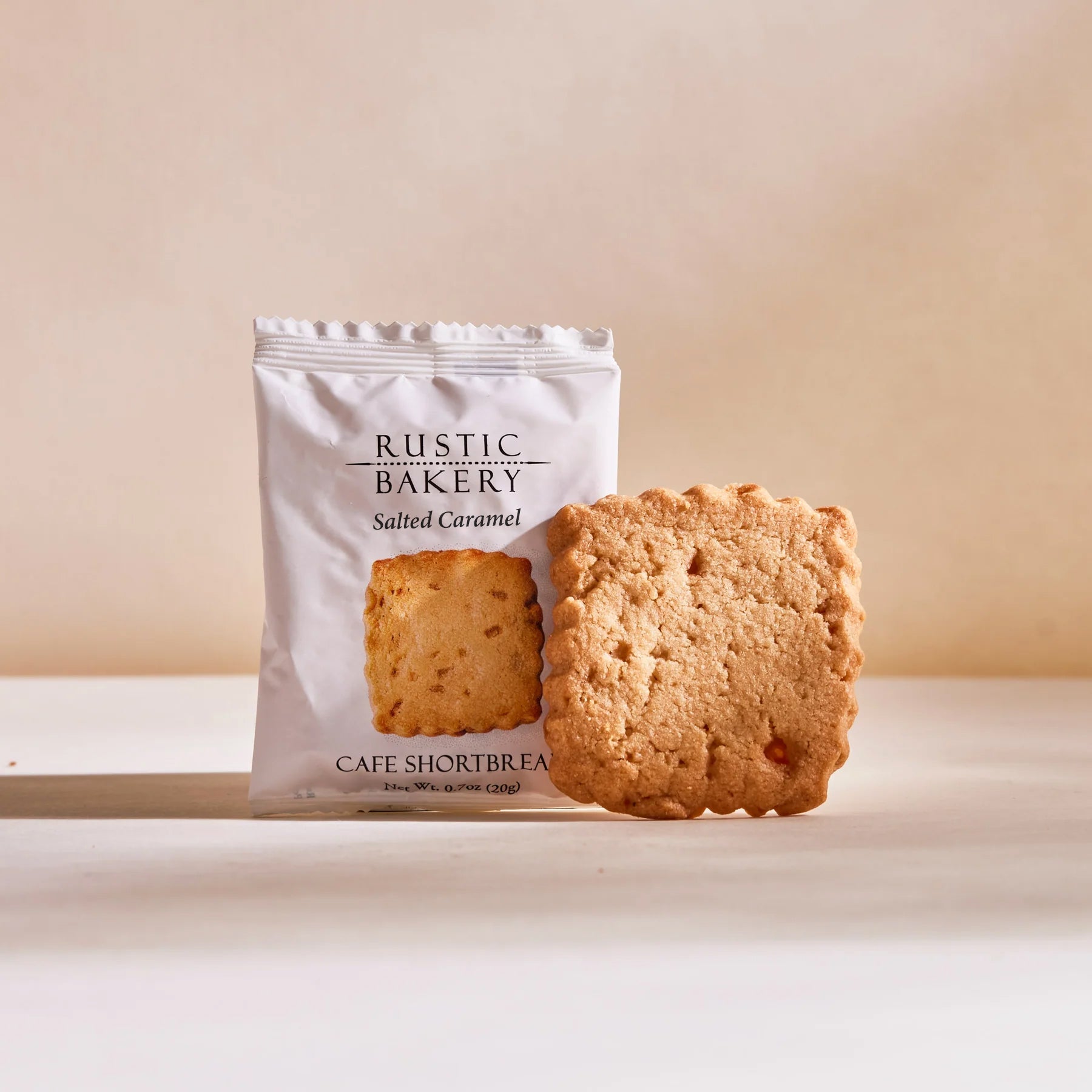 Rustic Bakery Salted Caramel Shortbread Cookie in its white packaging and outside of its packaging in a cream color background. 