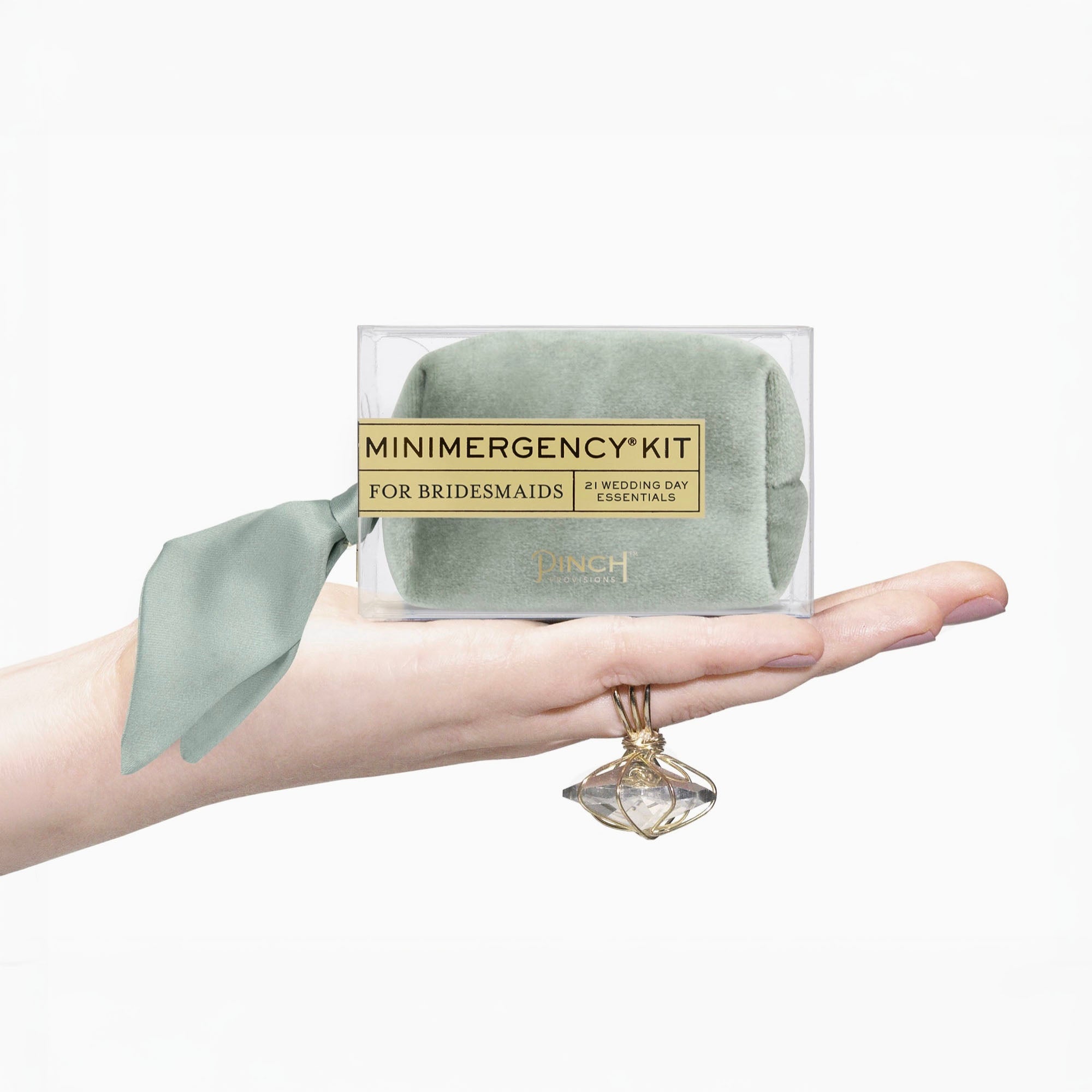 A hand holds a clear box containing a sage green velvet emergency kit for bridesmaids, with a matching sage green ribbon and a gold label. A gold ring is draped over the hand against a clean white background.