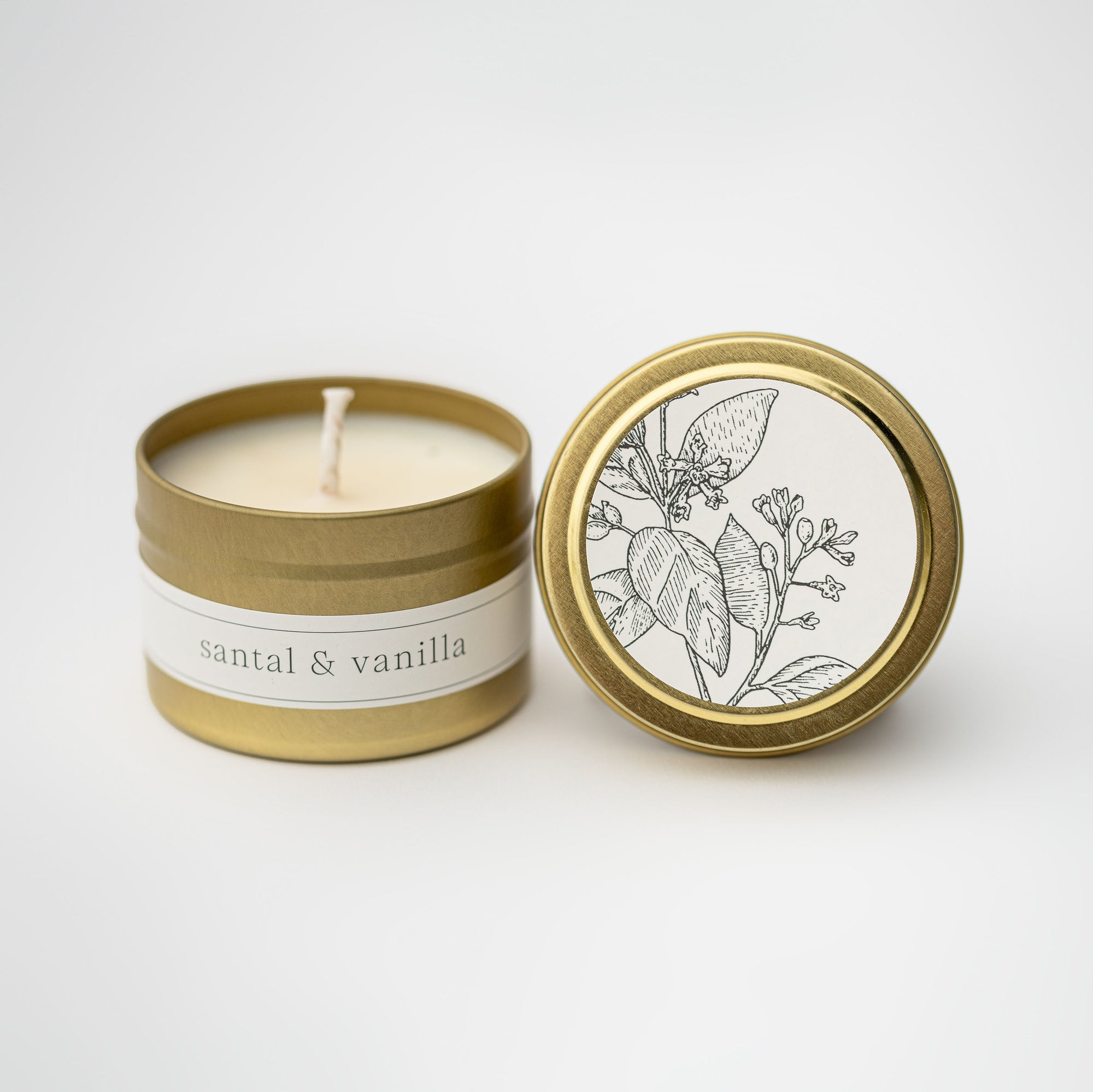 Santal and vanilla travel size candle featured in its gold tin and white label with flower illustration in black on lid and black lettering on tin on a white background. 