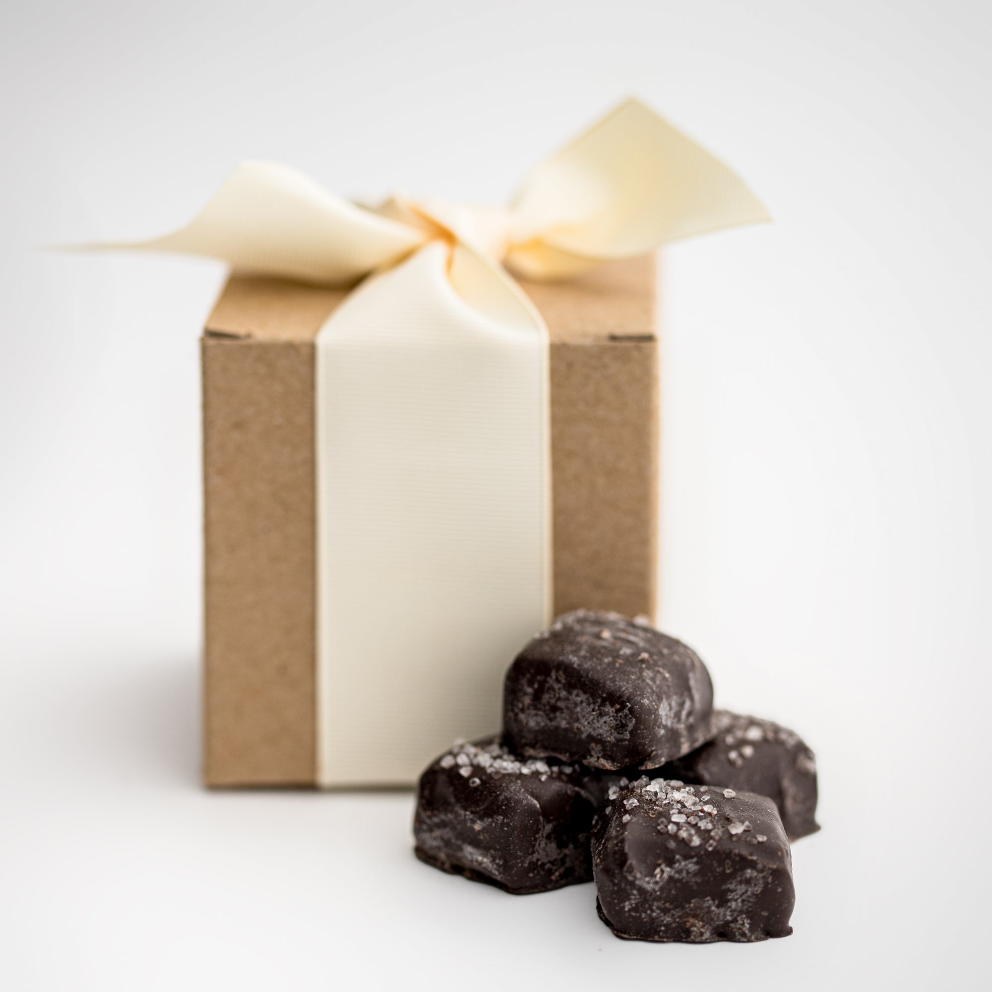Dark chocolate sea salt caramels are featured in the front, with four pieces displayed, and behind them, the light brown cardstock gift box they come in, wrapped with an oatmeal-colored ribbon.