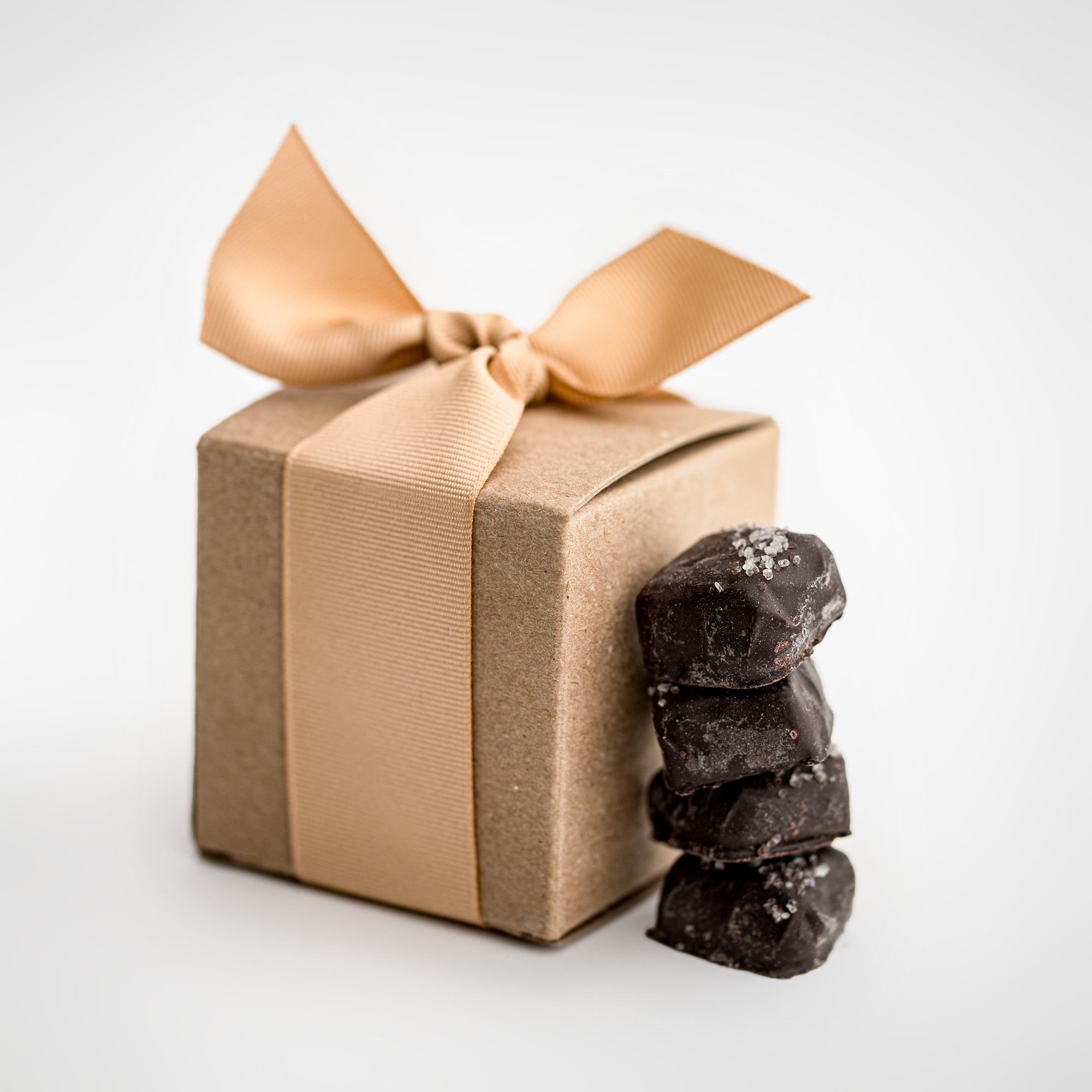 Dark chocolate sea salt caramels are featured in the front, with four pieces displayed, and behind them, the light brown cardstock gift box they come in, wrapped with an oatmeal-colored ribbon.