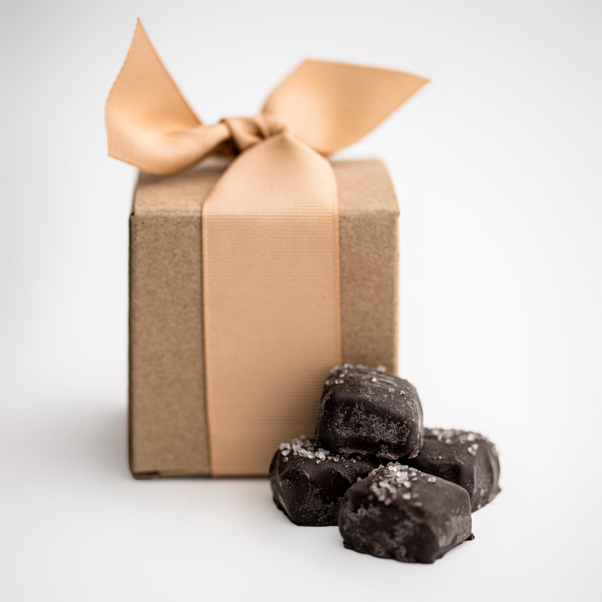 Dark chocolate sea salt caramels are featured in the front, with four pieces displayed, and behind them, the light brown cardstock gift box they come in, wrapped with an oatmeal-colored ribbon.