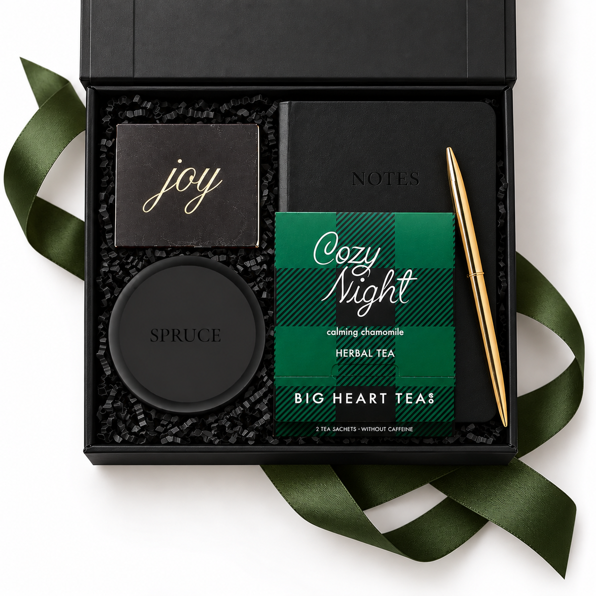 A luxury gift set in an open black box with black tissue paper, featuring a black soap bar with gold 'joy' script, a black round tin embossed with 'SPRUCE', a black notebook with 'NOTES' in gold, a green plaid 'Cozy Night' herbal tea packet, and a gold pen, all arranged with a dark green ribbon on a white background.