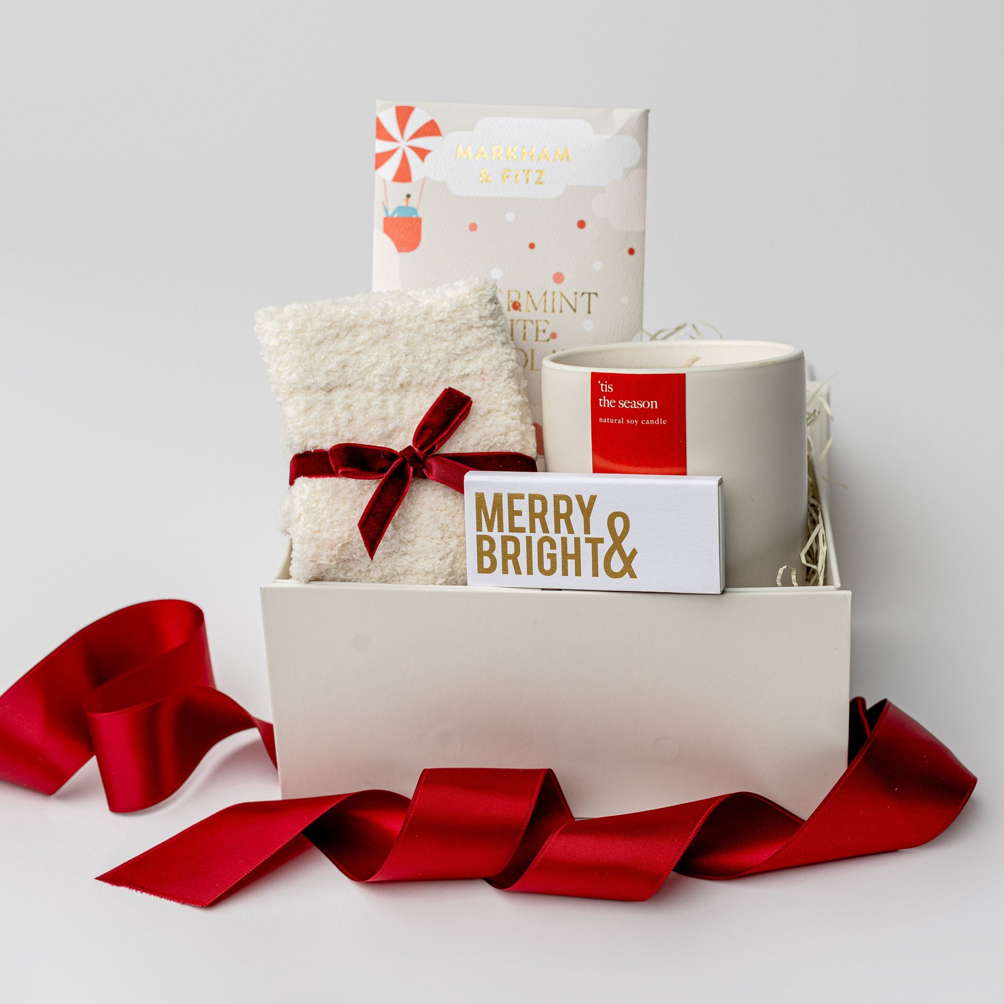 Secret Santa holiday gift box is featured a white small box with a red ribbon around it. inside of the box white on the left cozy white sock tied in red ribbon on the right this is the season candle with red label in the back peppermint  white chocolate bar and in the front merry & bright gold foil matchbox. All on a white background. 