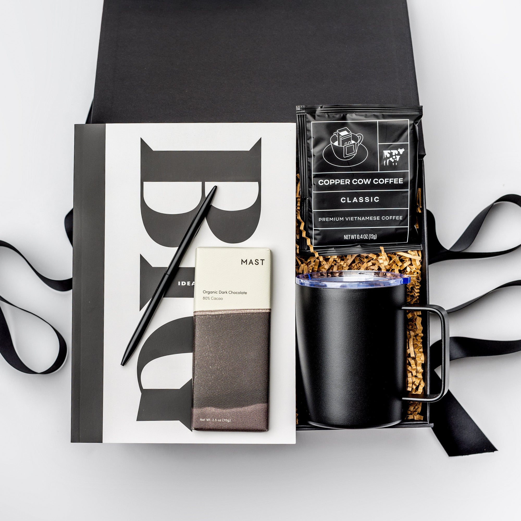 A top view of the Seize the Day Gift Box presented in a black gift box filled with brown shred and tied with a black ribbon, featuring a Big Idea sketchbook, a minimalist black bullet pen, a 12 oz. black vacuum insulated camp mug, a Copper Cow single-serve pour-over coffee, and a MAST organic dark chocolate bar (80% cacao), all arranged on a white background.