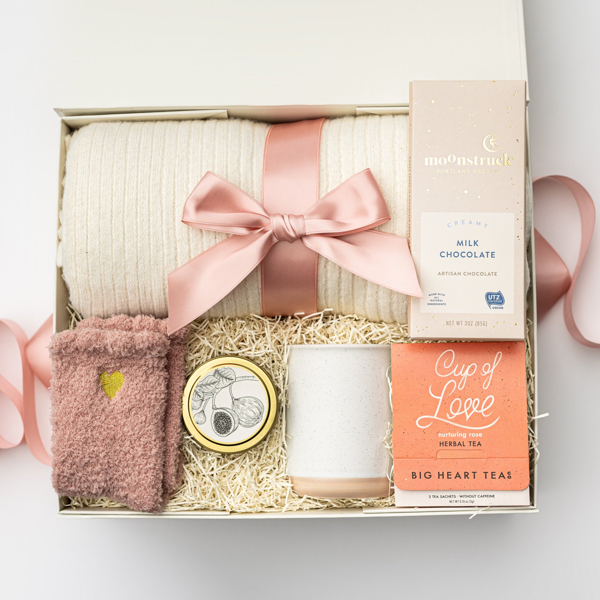 Top view of the “Sending Love” gift box displayed in a white box with a pink ribbon and ivory shred, containing a soft knit ivory throw blanket, a 12oz speckled ceramic mug, Cup of Love rose tea for two, a creamy milk chocolate bar, heart cozy socks, and a 4oz hand-poured Sweet Fig candle.