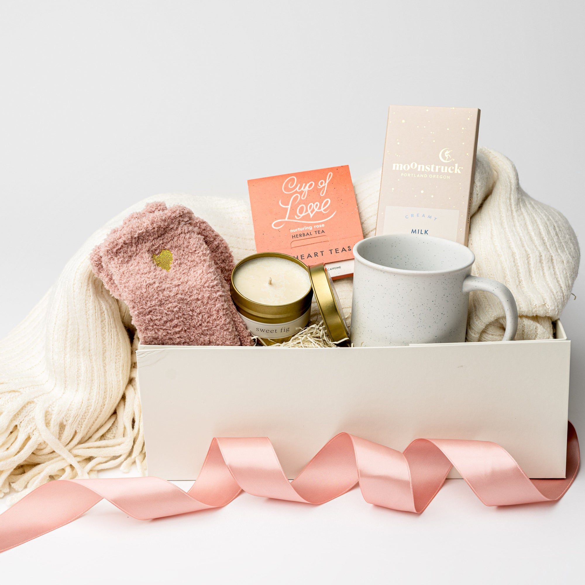Front view of the “Sending Love” gift box displayed in a white box with a pink ribbon and ivory shred, containing a soft knit ivory throw blanket, a 12oz speckled ceramic mug, Cup of Love rose tea for two, a creamy milk chocolate bar, heart cozy socks, and a 4oz hand-poured Sweet Fig candle.