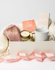 Front view of the “Sending Love” gift box displayed in a white box with a pink ribbon and ivory shred, containing a soft knit ivory throw blanket, a 12oz speckled ceramic mug, Cup of Love rose tea for two, a creamy milk chocolate bar, heart cozy socks, and a 4oz hand-poured Sweet Fig candle.