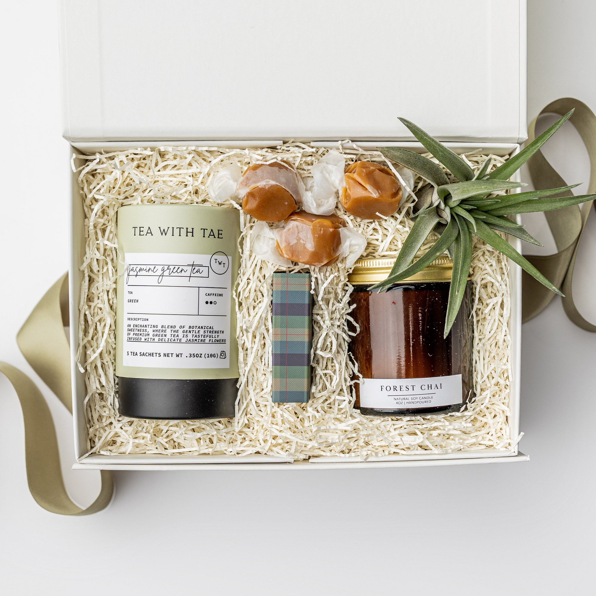 A white gift box with ivory paper shred and a green ribbon on a white background. Inside the box are a small live air plant with a care card, a mini tube of artisan jasmine green tea, a 4oz hand-poured soy candle in the scent Forest Chai, a matchbox of navy and green plaid-patterned matches, and three loose caramels — a thoughtful “Sending Thanks” gift box.