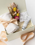 Serenity gift box by Foxblossom is featured. Inside the white box with ivory shred, a folded ivory-colored pashmina, tied with a delicate satin ribbon. To the left, a bouquet of dried flowers. On the right, a bar of white tea guest soap and a tube of shea hand cream from Santal Vanille. The box's ribbon spills out around the box set on a white background.