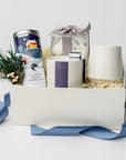 Silent Night Holiday Gift Box is featured open, revealing a white gift box with a blue ribbon in front of it. All arranged in front of white shred on the left side floral bundle, calming serene herbal tea, in the middle tea cakes, silent night candle with silver snuffer on top and on the left white speckled ceramic mug,. All on a white background.  