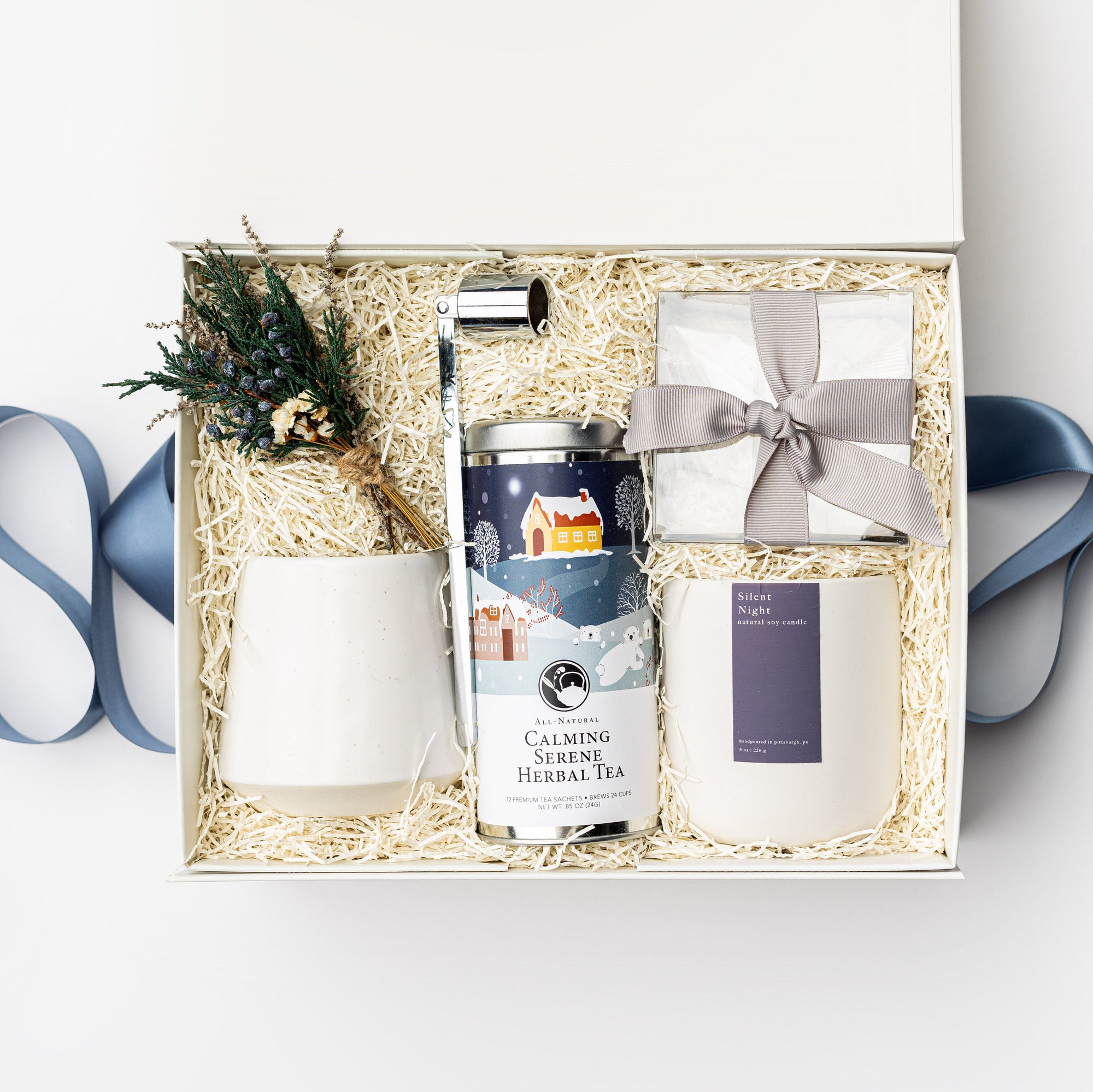 Silent Night Holiday Gift Box is featured open, a white gift box with a blue ribbon crossing underneath. Inside the box, arranged on top of white shred. On the left side, a floral bundle placed on top of a speckled white cup. In the middle, a silver candle snuffer and a calming serene herbal tea in a round silver tin with a Christmas-themed graphic label. On the right, a clear box containing tea cakes tied with a silver ribbon and a white ceramic “Silent Night” candle with a purple label, white background.