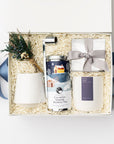 Silent Night Holiday Gift Box is featured open, a white gift box with a blue ribbon crossing underneath. Inside the box, arranged on top of white shred. On the left side, a floral bundle placed on top of a speckled white cup. In the middle, a silver candle snuffer and a calming serene herbal tea in a round silver tin with a Christmas-themed graphic label. On the right, a clear box containing tea cakes tied with a silver ribbon and a white ceramic “Silent Night” candle with a purple label, white background.