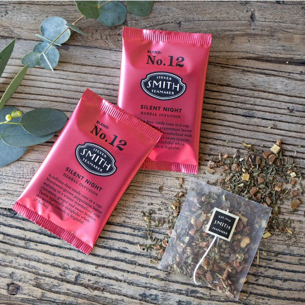 Two silent night tea bags are featured red with black lettering on a wooden background. On the right side one of the sachets showing the tea has a black label with white lettering. A tree branch  on the left. 