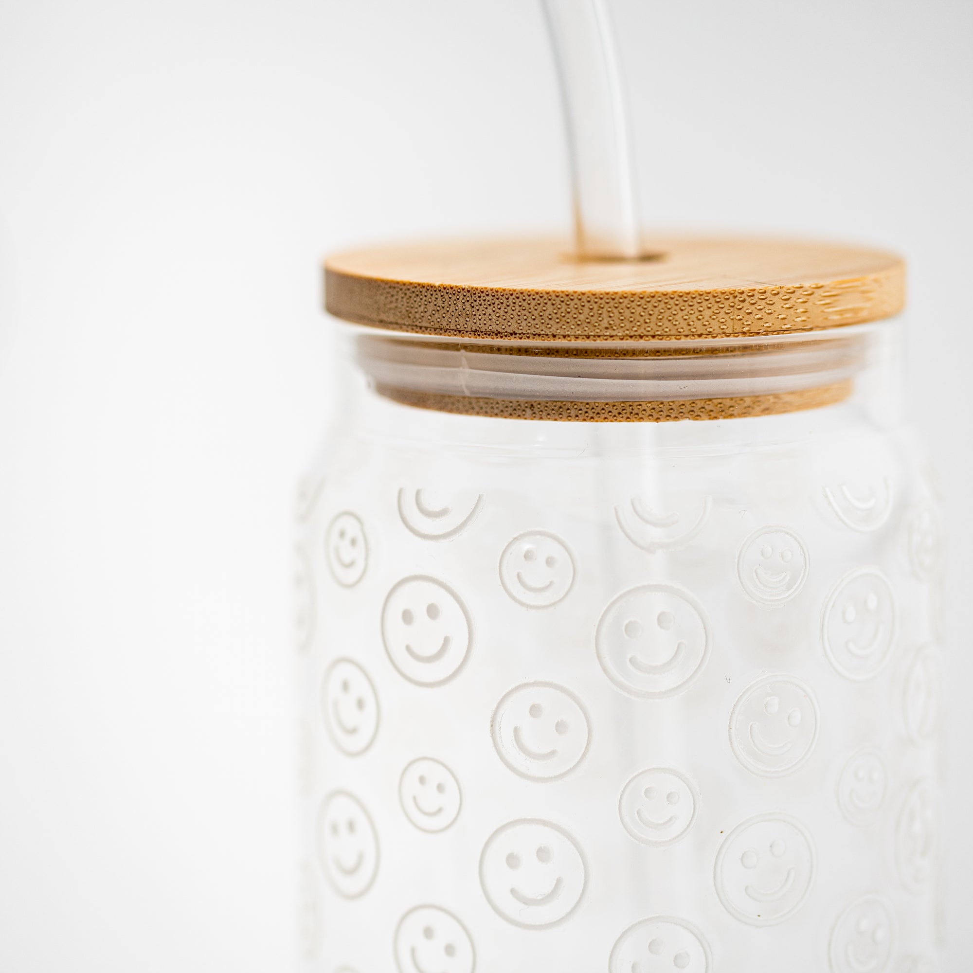 A smiley print glass cup close-up image is shown, in its glass cup with a printed white smiley with a wooden lid and a glass straw on a white background.