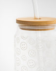 A smiley print glass cup close-up image is shown, in its glass cup with a printed white smiley with a wooden lid and a glass straw on a white background.