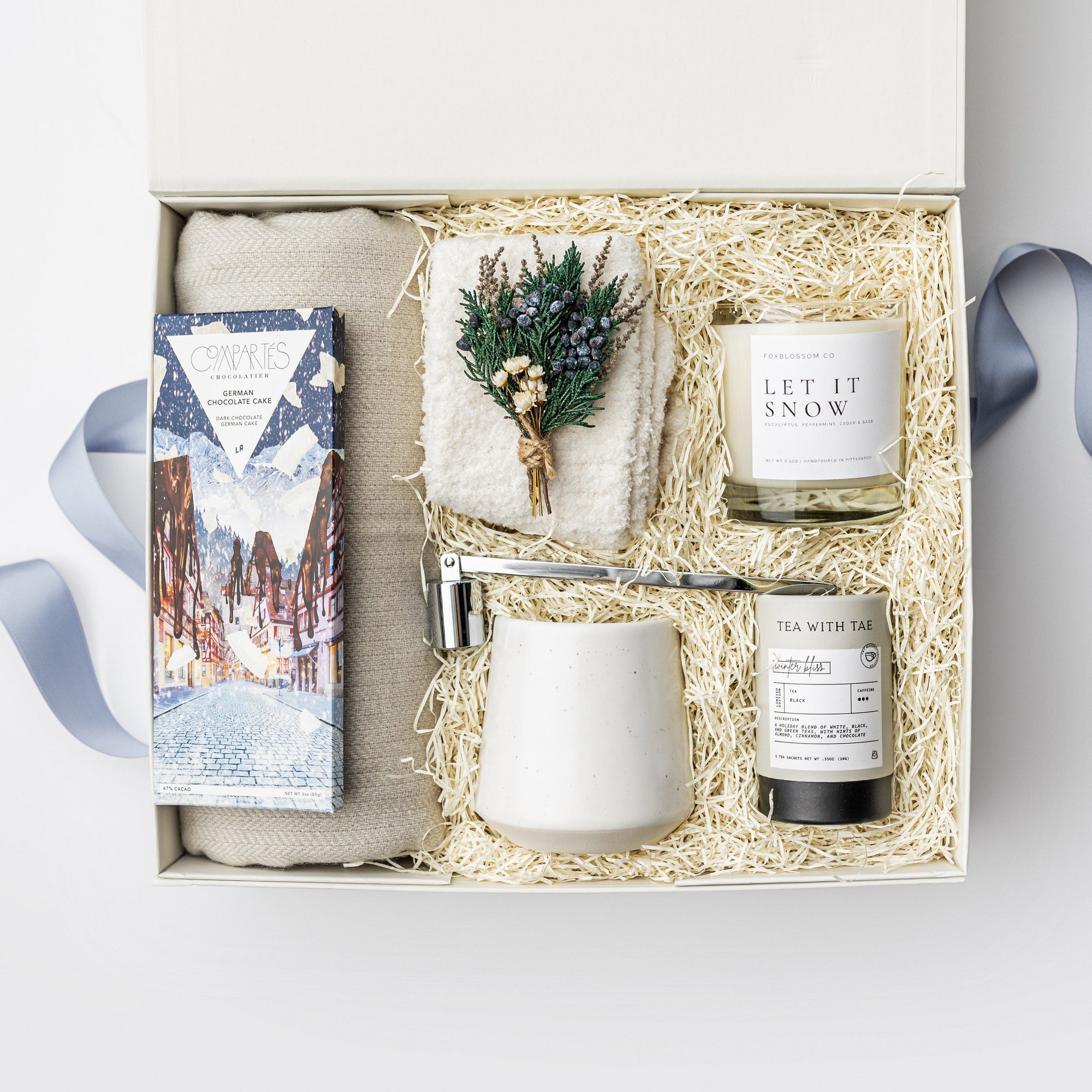 A white gift box on a white background, filled with ivory paper shred. Inside the box: a light-gray wool-blend throw blanket (50 × 60″), a pair of unisex cozy cloud socks, a mini tube of Winter Bliss tea, a 10 oz speckled ceramic mug, a Compartes German Chocolate Cake bar, a 9 oz hand-poured soy candle labeled “Let It Snow,” and a silver metal candle snuffer — a snug, holiday “Snow Day” gift set.