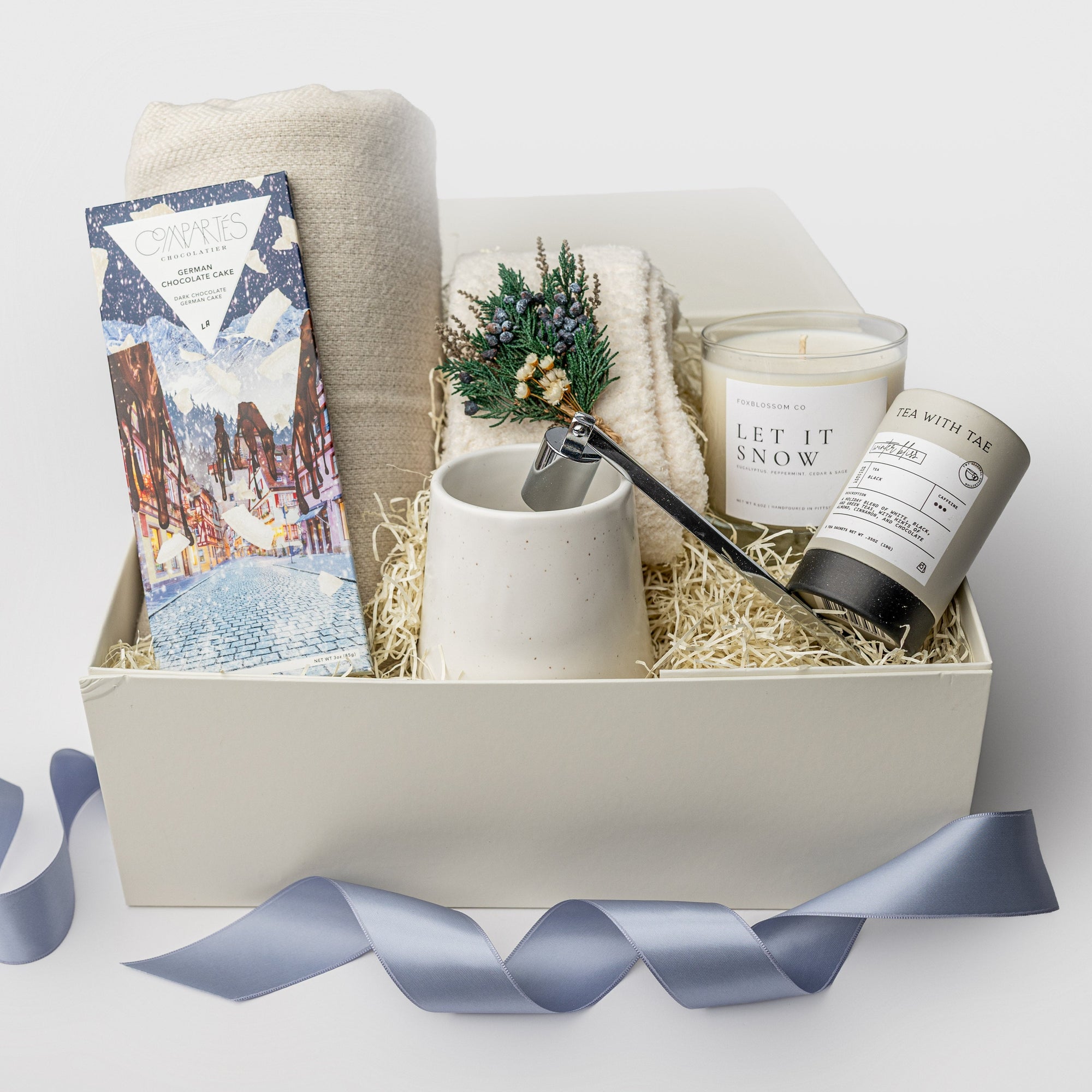 A white gift box on a white background, filled with ivory paper shreds. Inside the box: a light-gray wool-blend throw blanket (50 × 60″), a pair of unisex cozy cloud socks, a mini tube of Winter Bliss tea, a 10 oz speckled ceramic mug, a Compartes German Chocolate Cake bar, a 9 oz hand-poured soy candle labeled “Let It Snow,” and a silver metal candle snuffer — a snug, holiday “Snow Day” gift set.