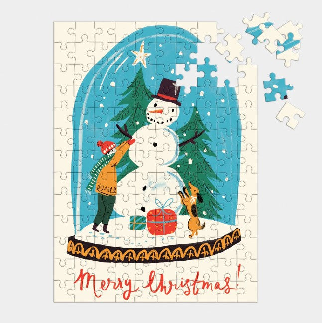 Snow globe mini puzzle is featured almost completely done showing a snow globe illustration with merry Christmas in red on the bottom with some pieces not assembled in the puzzle on the right corner on a grayish background. 